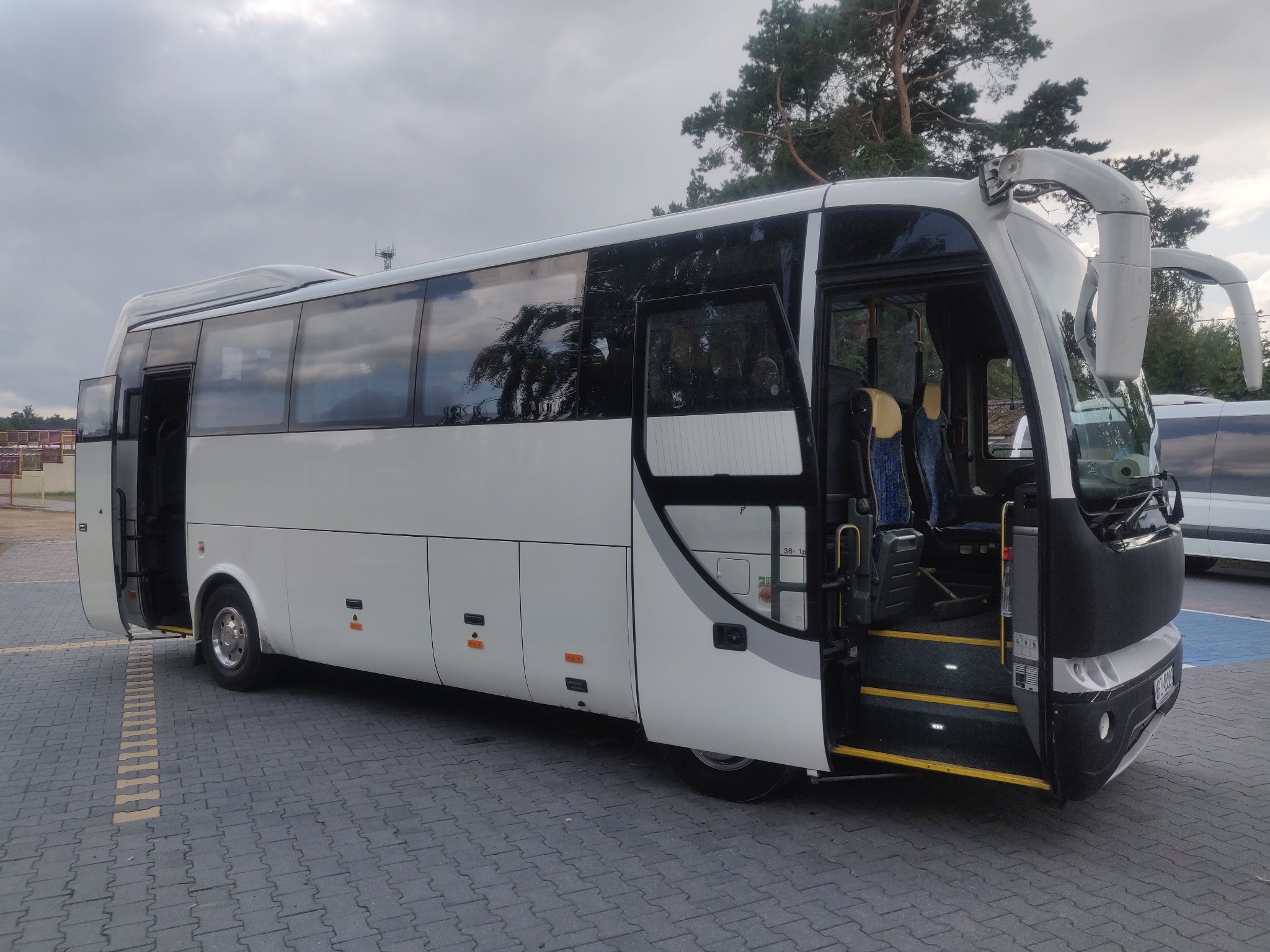 Biały bus z otwartymi drzwiami na parkingu z kostki brukowej, widoczne schodki wejściowe z żółtymi krawędziami i częściowo widoczne wnętrze z niebieskimi siedzeniami.