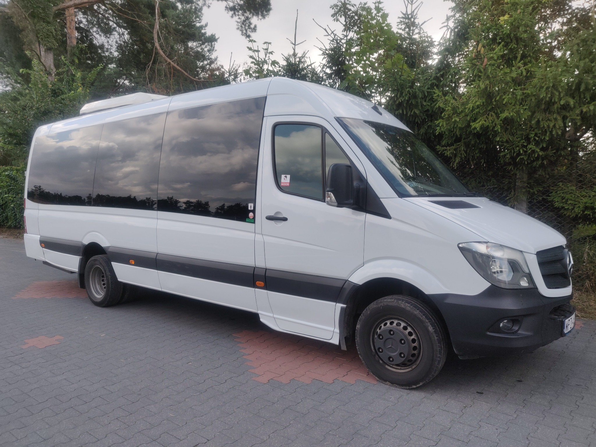 Biały, długi bus z czarnymi szybami i klimatyzacją na dachu, zaparkowany na szarej kostce brukowej, na tle zielonych drzew.