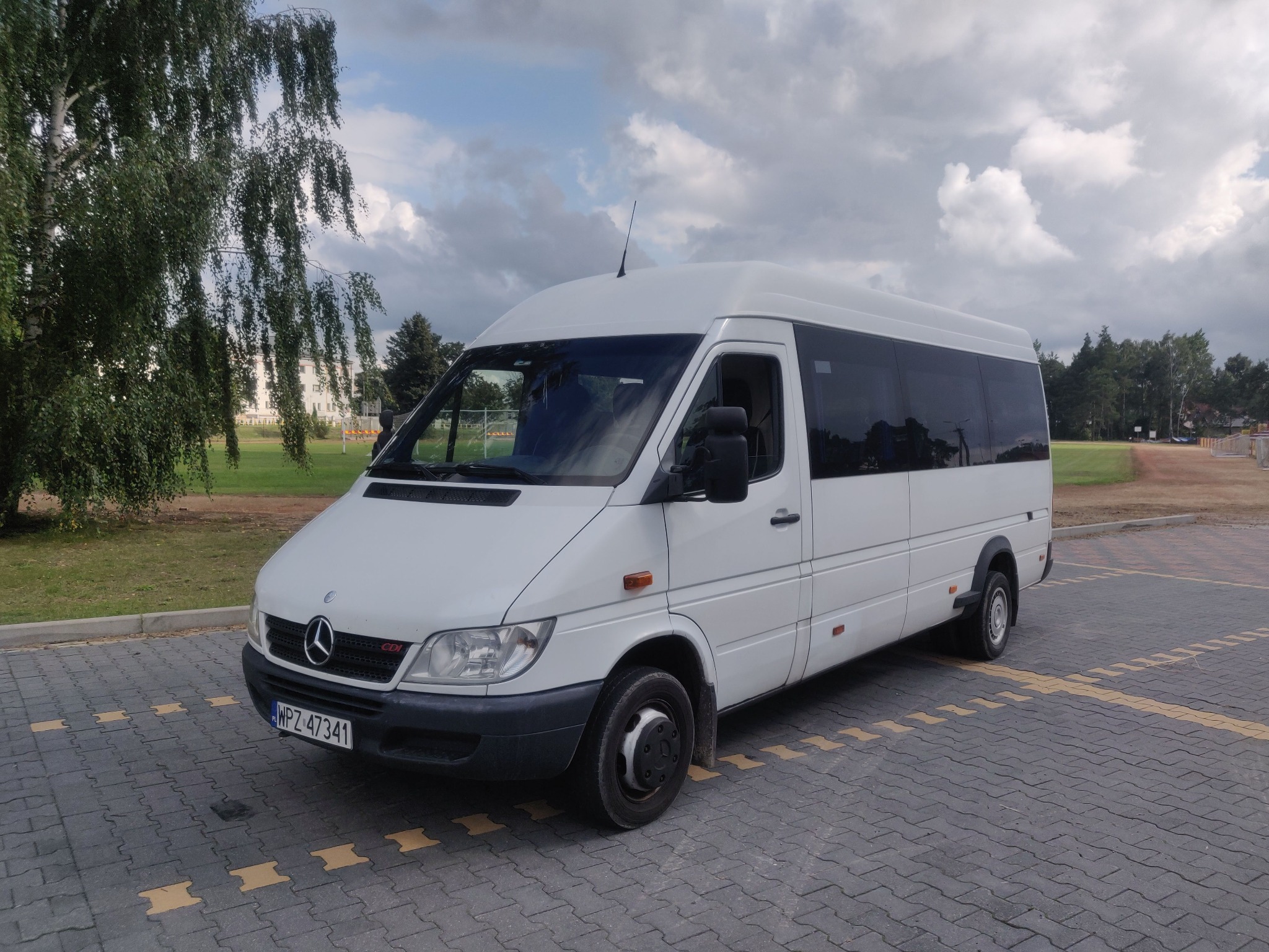 Biały bus Mercedes-Benz Sprinter z wysokim dachem, zaparkowany na brukowanej powierzchni z żółtymi oznaczeniami, na tle pochmurnego nieba i drzew.