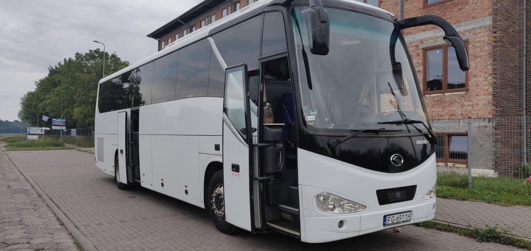 Biały autokar z otwartymi drzwiami na parkingu, widoczny numer rejestracyjny i budynek z cegły w tle.