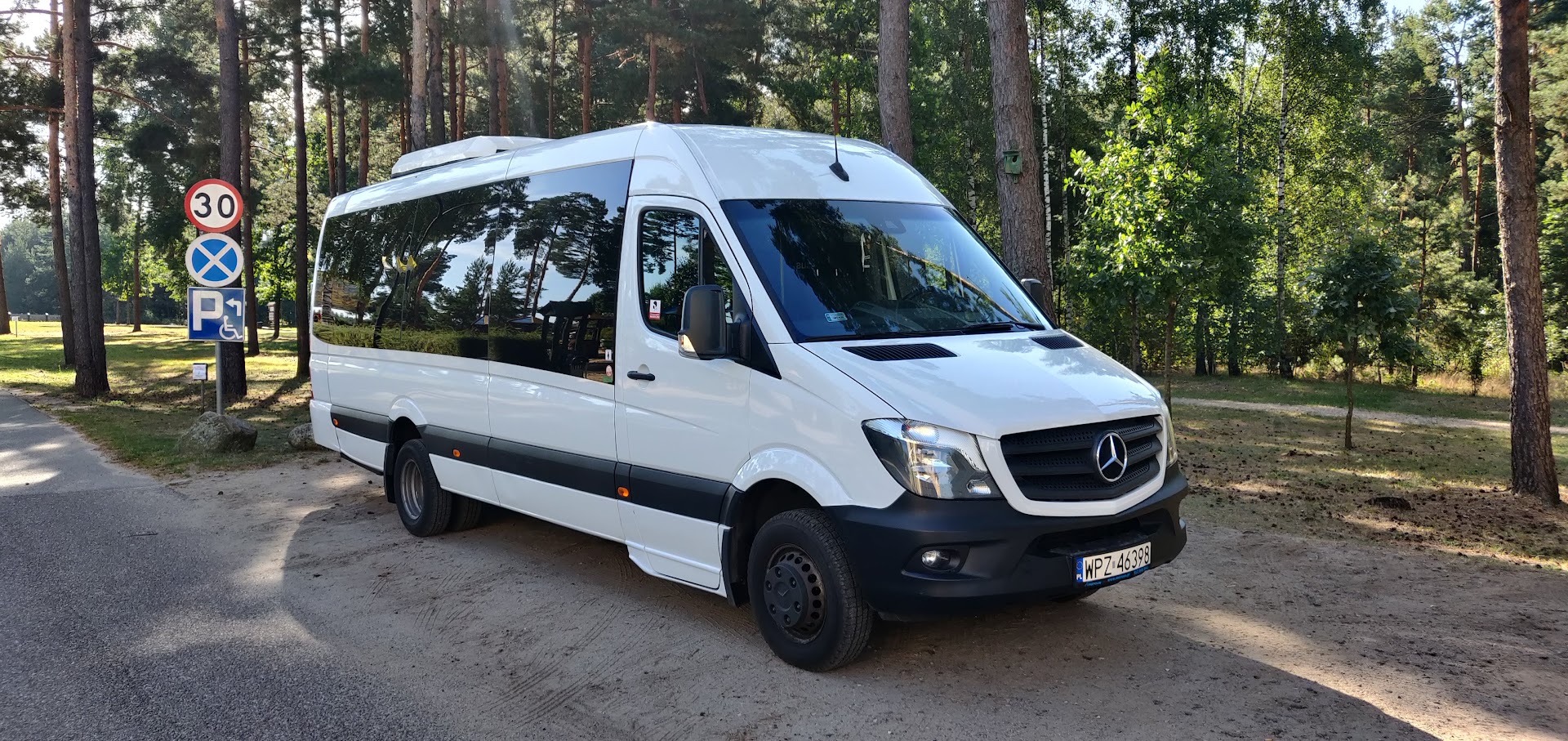 Biały bus Mercedes-Benz Sprinter z przyciemnianymi szybami, zaparkowany na leśnym parkingu obok znaku drogowego z ograniczeniem prędkości do 30 km/h i symbolem parkingu dla osób niepełnosprawnych.