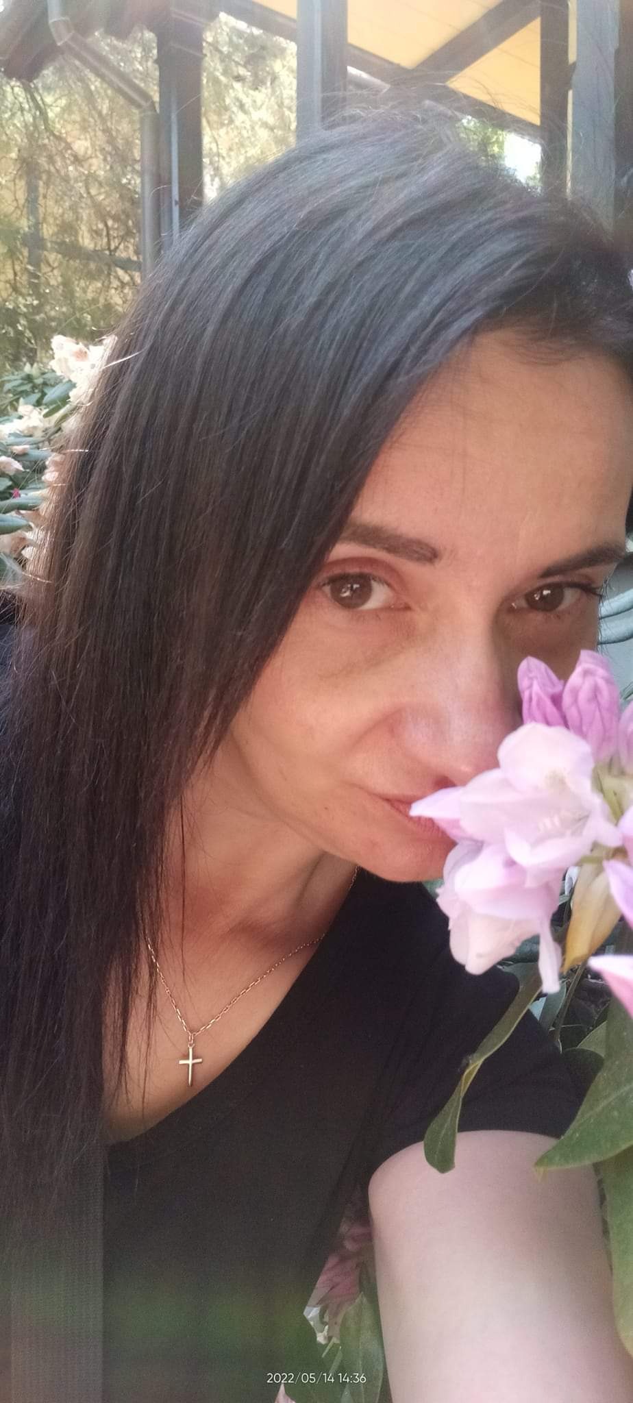 Portret kobiety z ciemnymi włosami i złotym naszyjnikiem z krzyżykiem, wąchającej jasnoróżowe kwiaty rododendronu w ogrodzie.