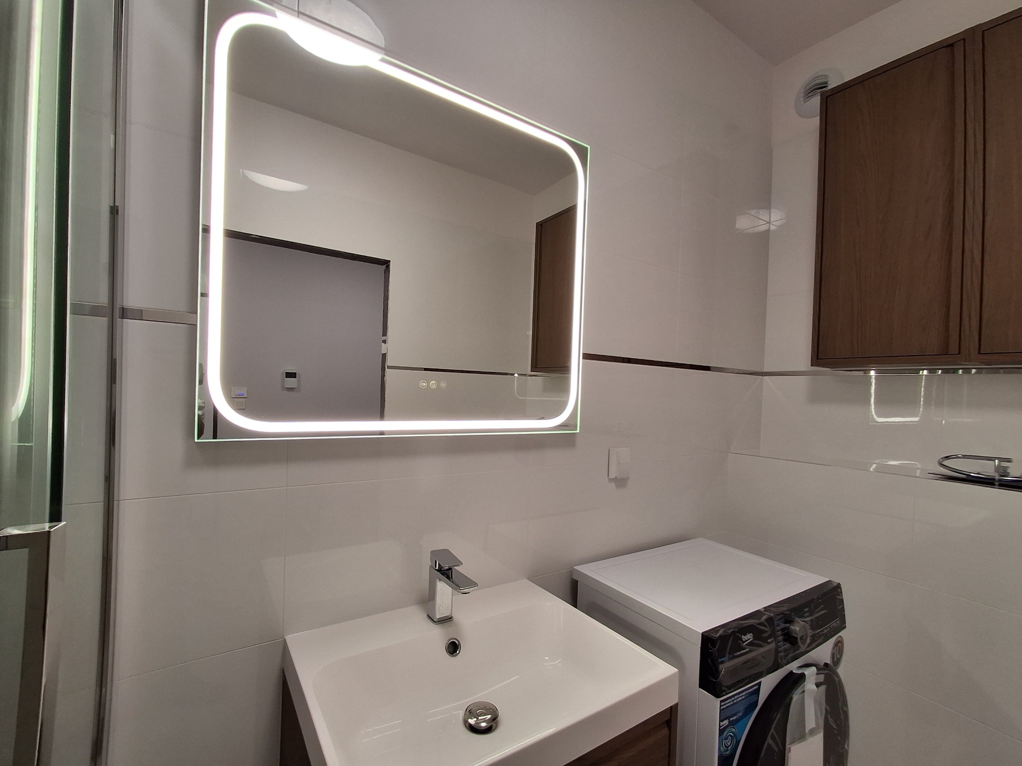 Nowoczesna łazienka z podświetlanym lustrem, umywalką nablatową, pralką i brązowymi szafkami wiszącymi na białych płytkach.