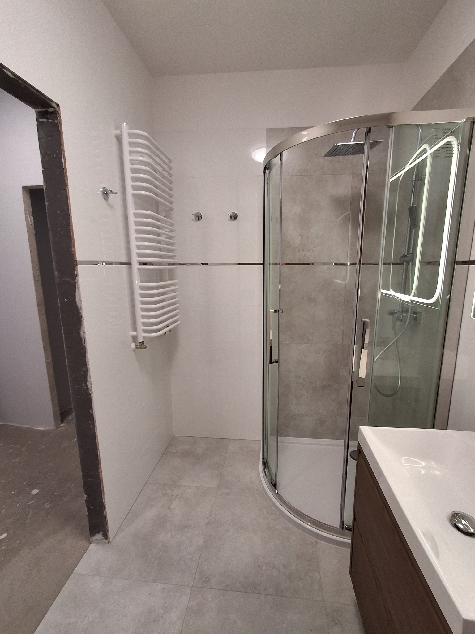 Wykończona łazienka z kabiną prysznicową typu walk-in z szarymi płytkami, grzejnikiem drabinkowym i umywalką z szafką w odcieniach brązu, widok na nieukończone przejście do innego pomieszczenia.