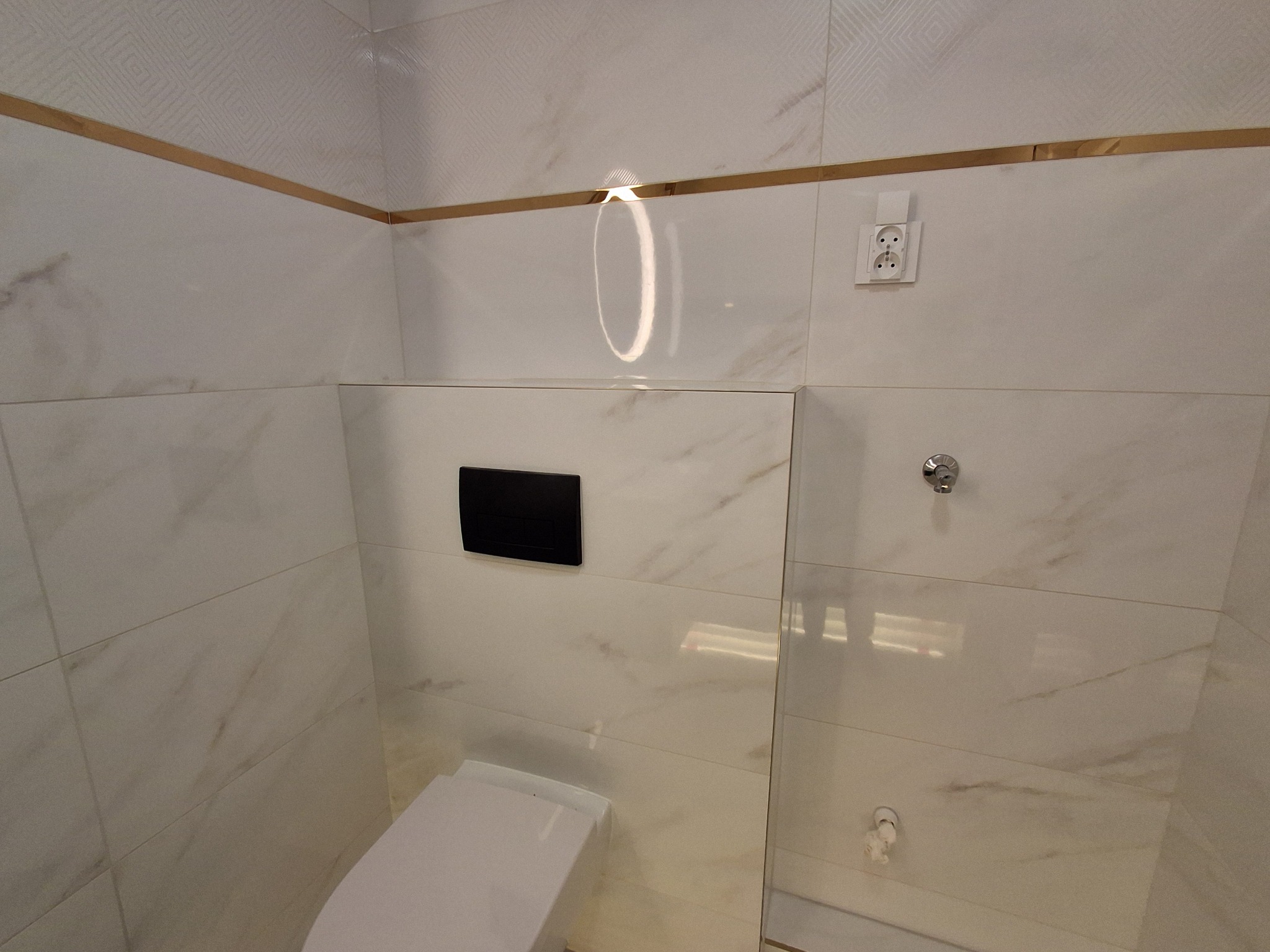 Wykończona toaleta z białymi płytkami imitującymi marmur, złotymi listwami dekoracyjnymi, czarnym panelem sterującym spłuczką i gniazdkiem elektrycznym na ścianie.