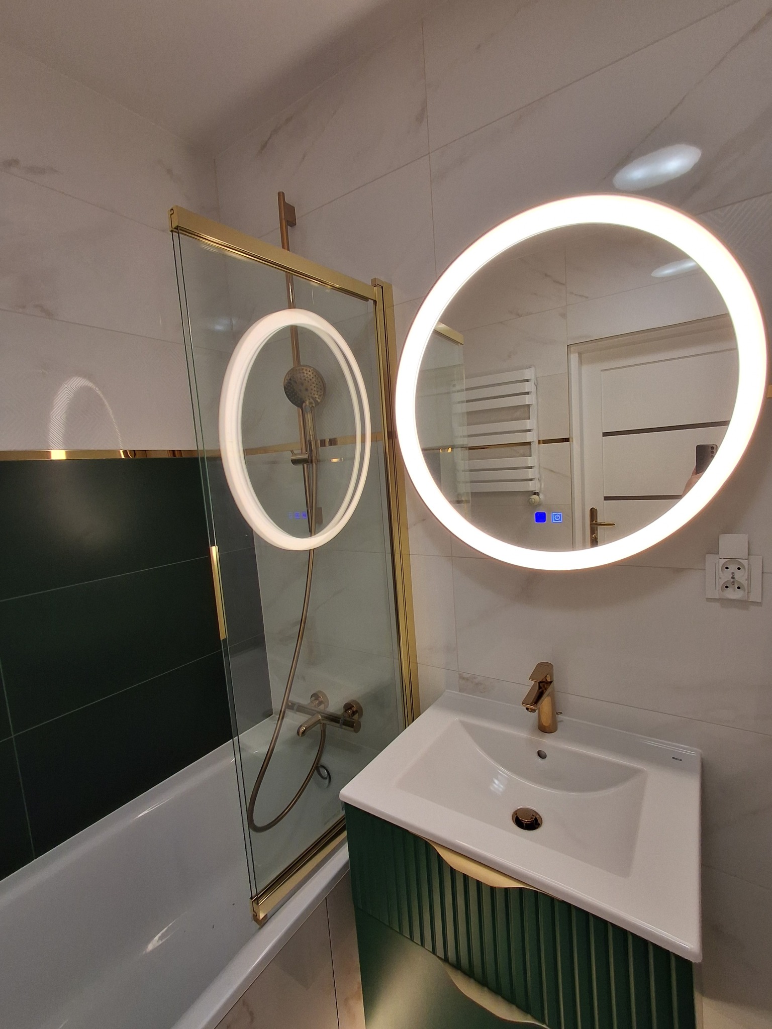 Nowoczesna łazienka z wanną za szklanym parawanem, umywalką na zielonej szafce z frezowanymi drzwiami i okrągłym lustrem z podświetleniem LED.