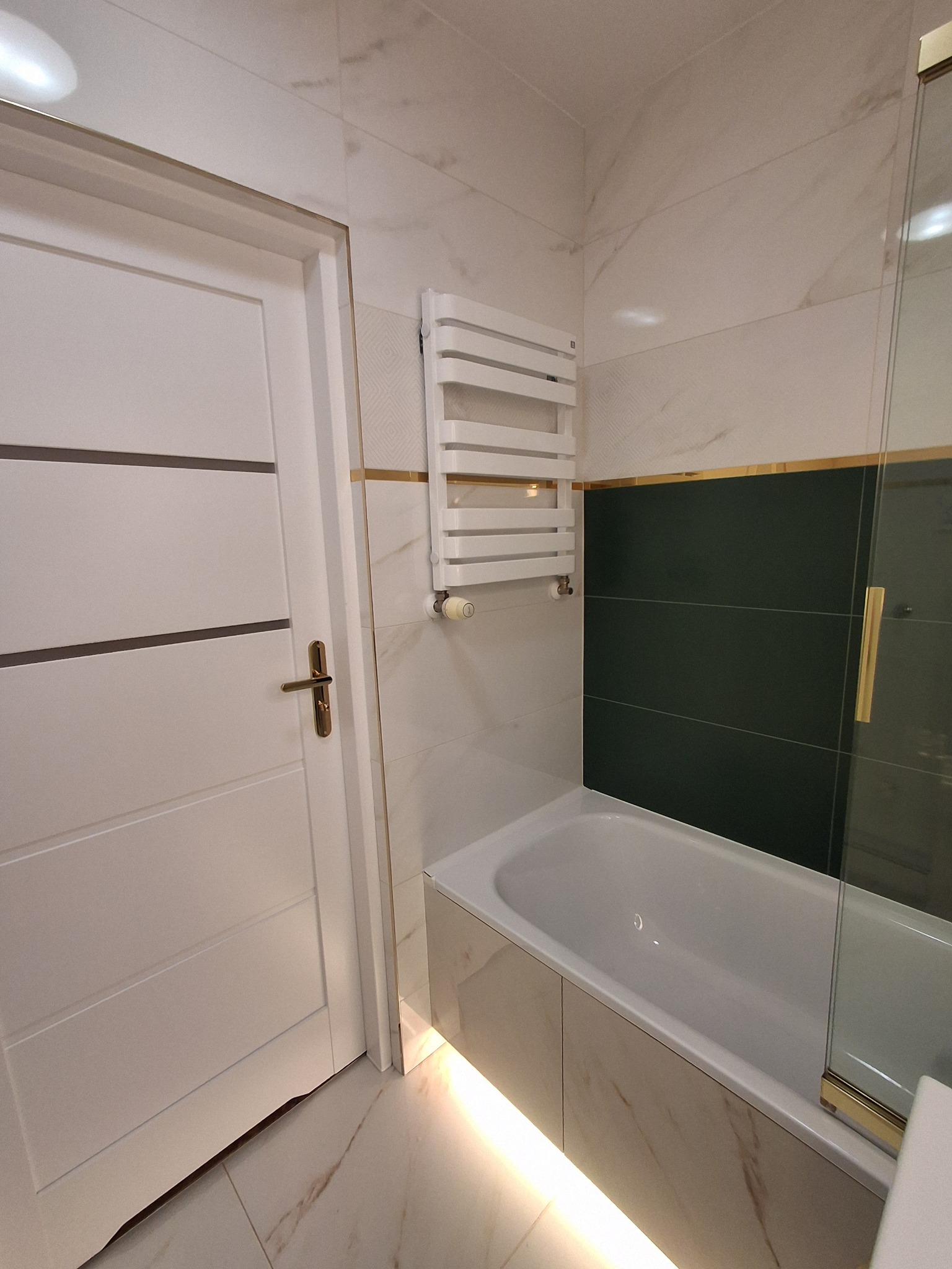 Nowoczesna łazienka z białymi marmurkowymi płytkami, zielonymi akcentami, białą wanną obudowaną płytkami, białym grzejnikiem drabinkowym i drzwiami z poziomymi frezami oraz złotymi detalami.