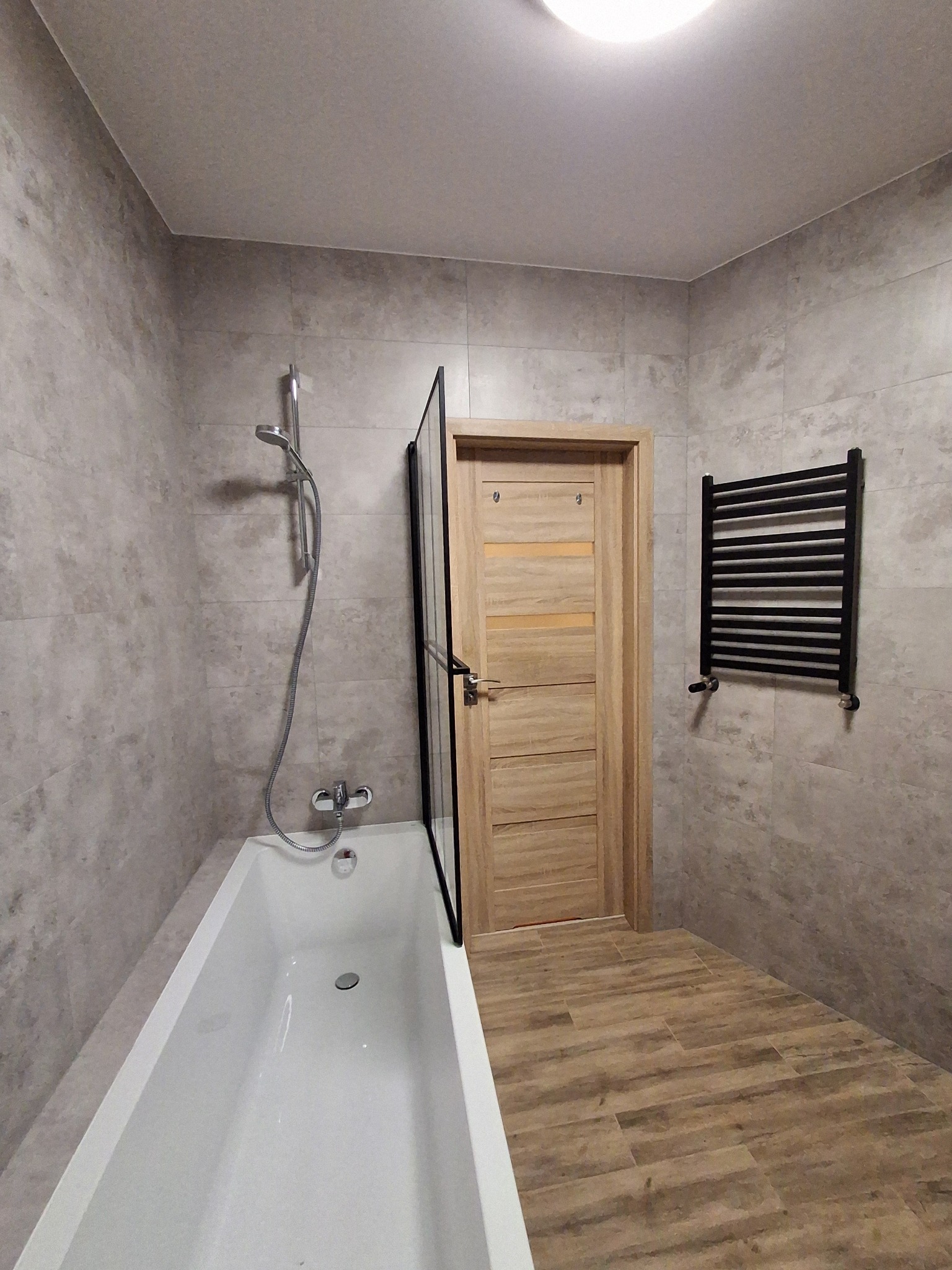 Nowoczesna łazienka z wanną, szklanym parawanem, grzejnikiem drabinkowym i płytkami imitującymi beton na ścianach oraz drewnopodobnymi na podłodze, oświetlona górnym światłem.