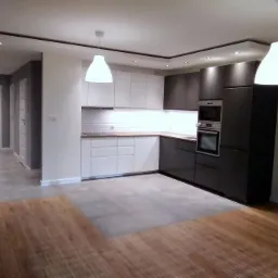 Nowoczesna, otwarta kuchnia z białymi i grafitowymi szafkami, podświetlanym blatem, białymi płytkami na ścianie i drewnianą podłogą przechodzącą w szare płytki.