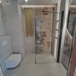 Nowoczesna łazienka z płytek imitujących kamień, z kabiną prysznicową typu walk-in z czarnymi profilami, podwieszaną toaletą i dekoracyjnym, złotym grzejnikiem drabinkowym.