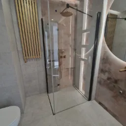 Nowoczesna kabina prysznicowa z czarnymi profilami i złotą deszczownicą, na tle marmurowej ściany z podświetleniem LED, obok złoty grzejnik drabinkowy i toaleta.