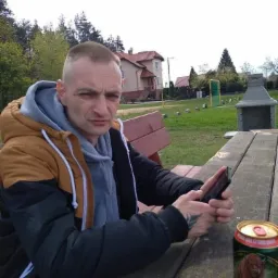 Mężczyzna w kurtce siedzi przy drewnianym stole na zewnątrz, trzyma telefon w dłoni, obok stoi puszka piwa Żubr i leży batonik.