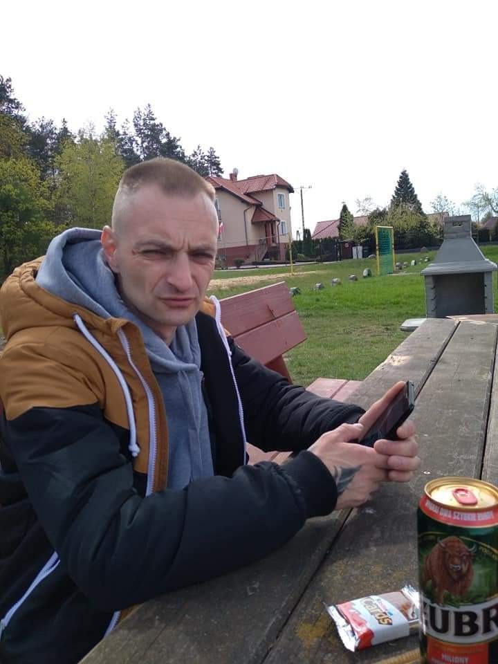 Mężczyzna w kurtce siedzi przy drewnianym stole na zewnątrz, trzyma telefon w dłoni, obok stoi puszka piwa Żubr i leży batonik.