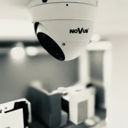 Kamera kopułkowa marki Novus zamontowana na suficie w pomieszczeniu, widok z dołu w czerni i bieli, z rozmytym tłem.
