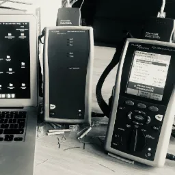 Czarno-białe zdjęcie miejsca pracy instalatora sieciowego: laptop, tester kabli Fluke DTX-1800 z podłączonym adapterem, fragmenty przewodów i złączki RJ45 na biurku.