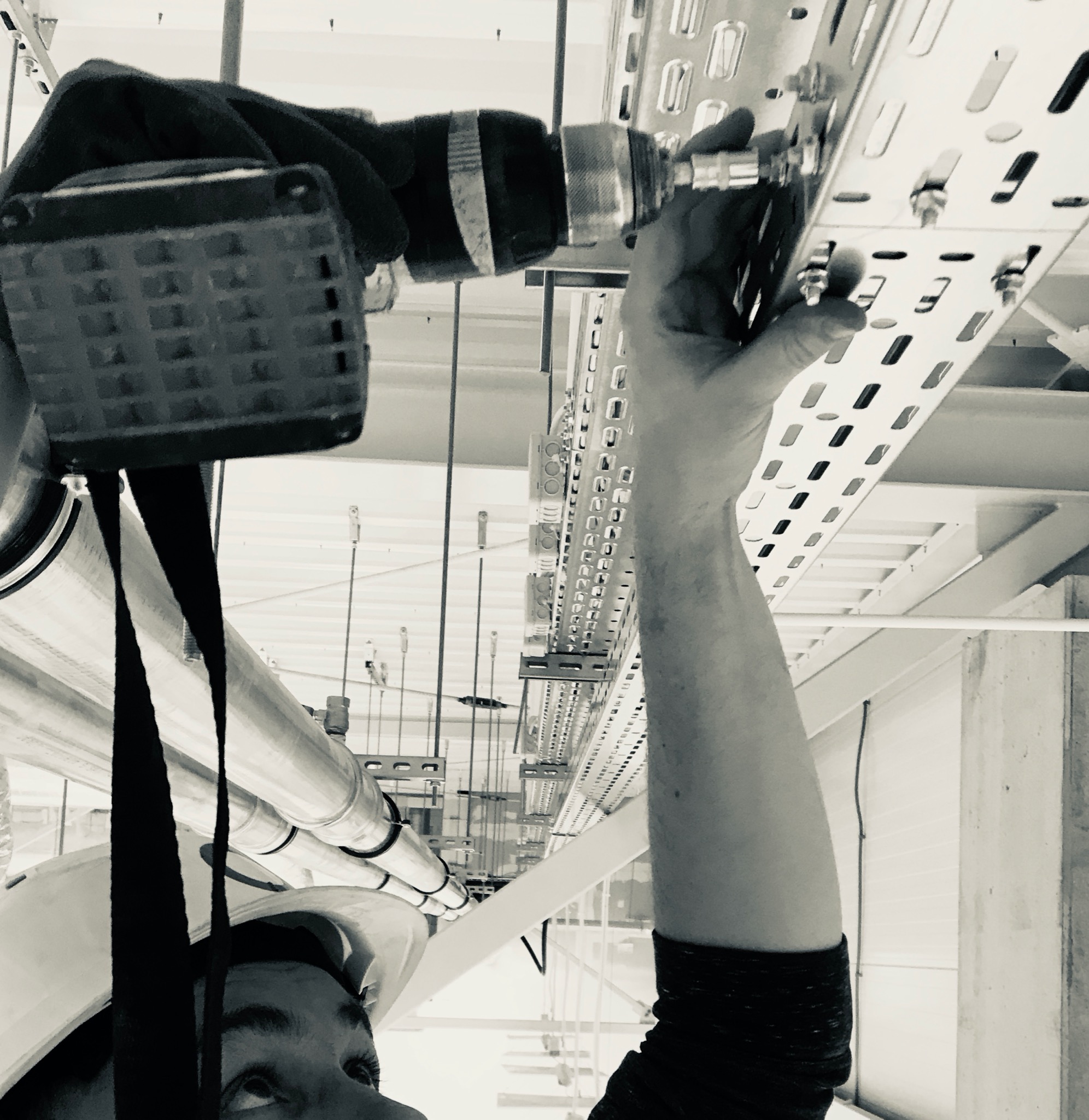 Czarno-białe zdjęcie instalatora w kasku, montującego metalową rurę do perforowanej szyny montażowej pod sufitem, widok z dołu.