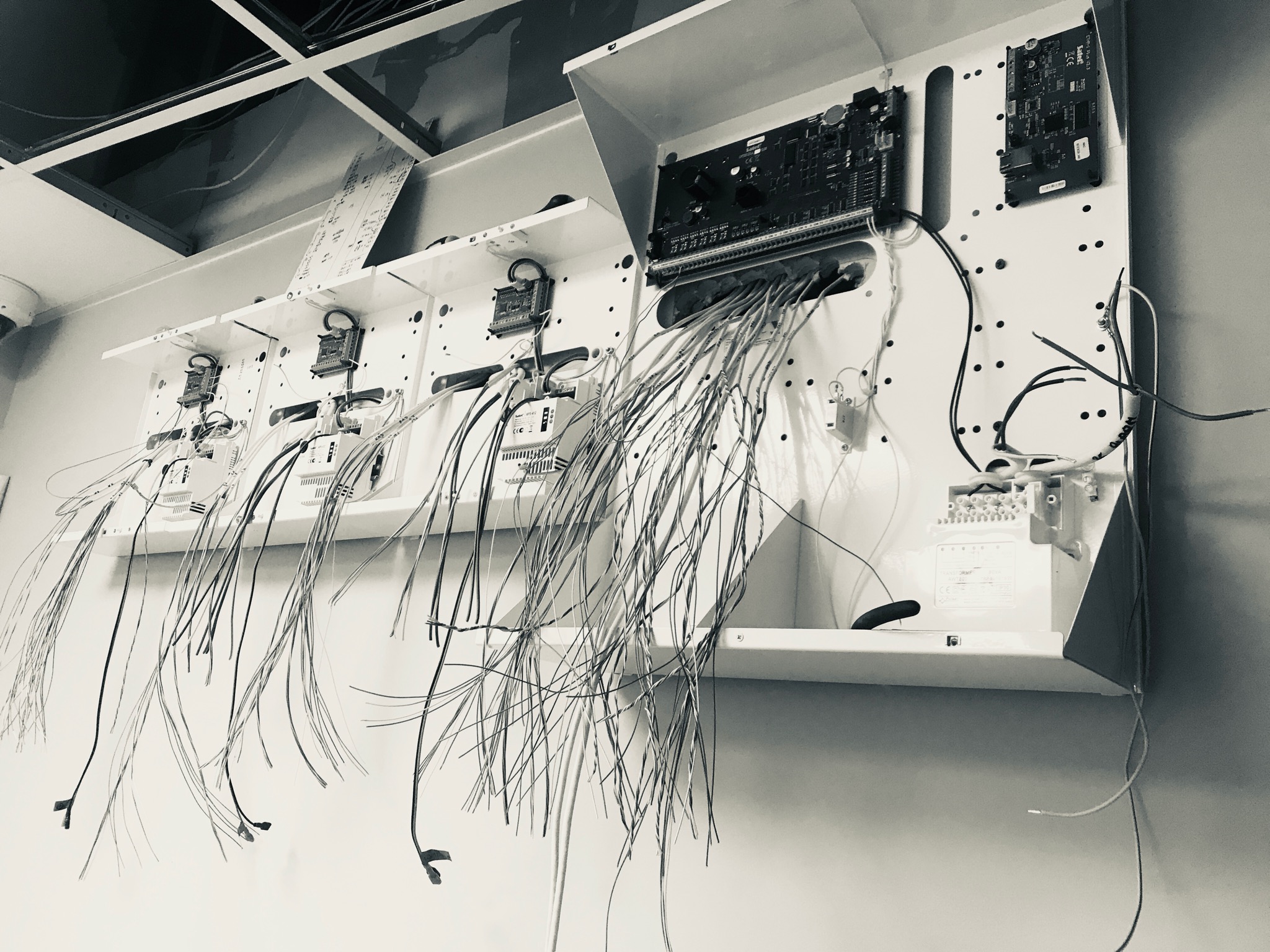 Czarno-białe zdjęcie w trakcie instalacji centrali alarmowej z widocznymi licznymi przewodami zwisającymi spod obudowy, zamontowanej na ścianie w pomieszczeniu z podwieszanym sufitem.