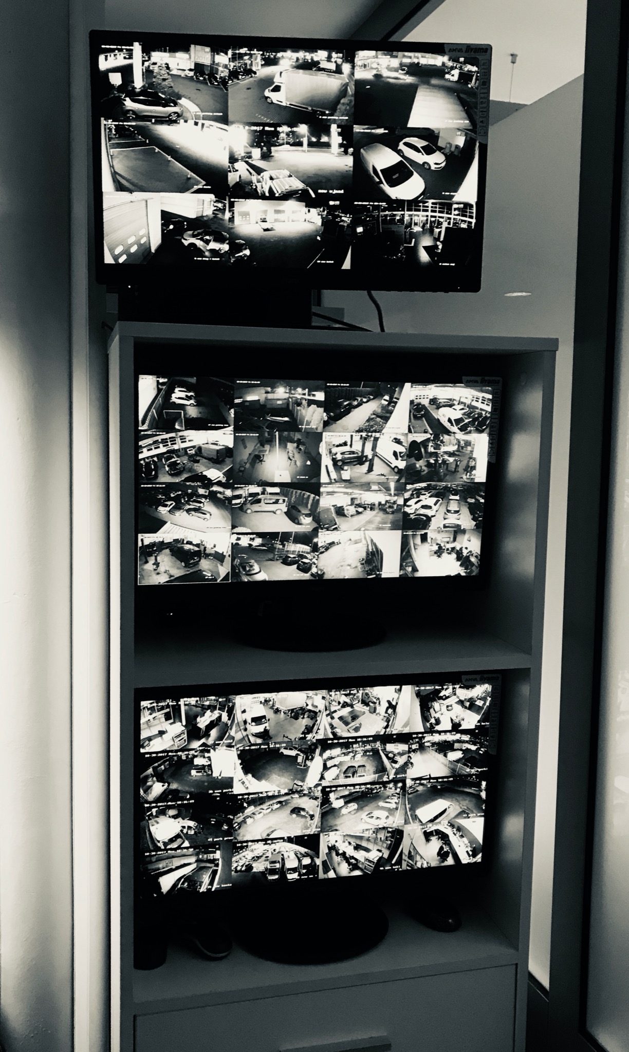 Trzy ekrany monitoringu CCTV umieszczone jeden nad drugim w szarym regale, prezentujące obraz z wielu kamer w czarno-białej tonacji, ukazujące parkingi i wjazdy do budynków.