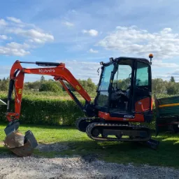 Pomarańczowa minikoparka Kubota KX027-4 na żwirowym podłożu w Goraju, z widocznym lemieszem i łyżką, na tle zielonego trawnika i błękitnego nieba z chmurami.