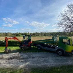 Pomarańczowa minikoparka Kubota obok zielono-żółtej wywrotki na żwirowej drodze, w tle pole i błękitne niebo z chmurami.