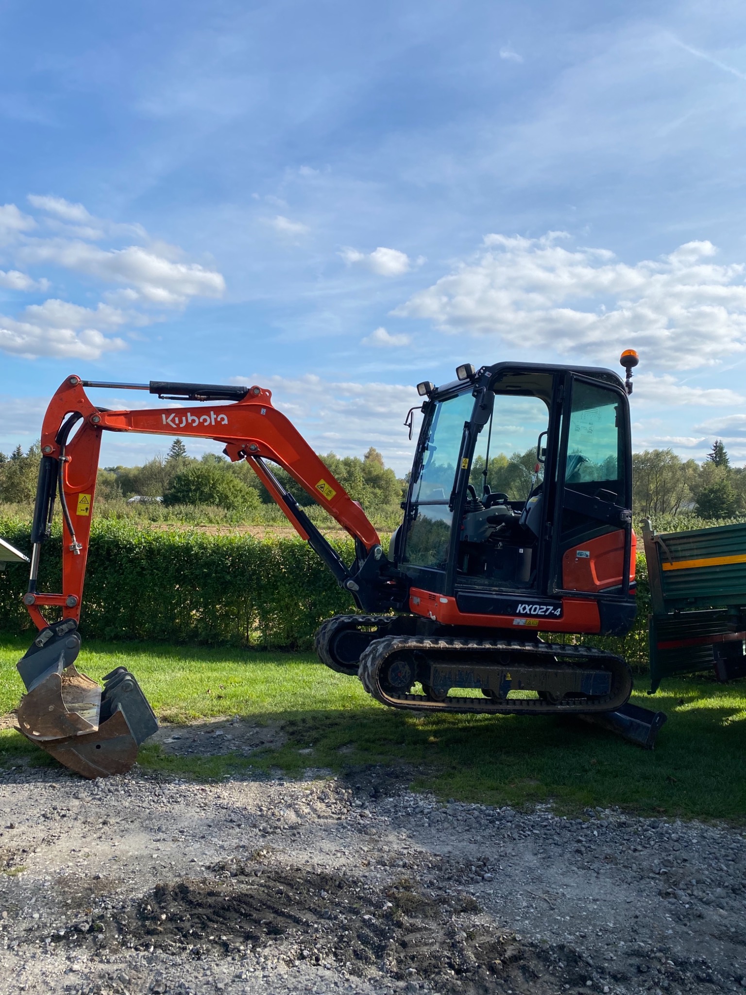 Pomarańczowa minikoparka Kubota KX027-4 na żwirowym podłożu w Goraju, z widocznym lemieszem i łyżką, na tle zielonego trawnika i błękitnego nieba z chmurami.