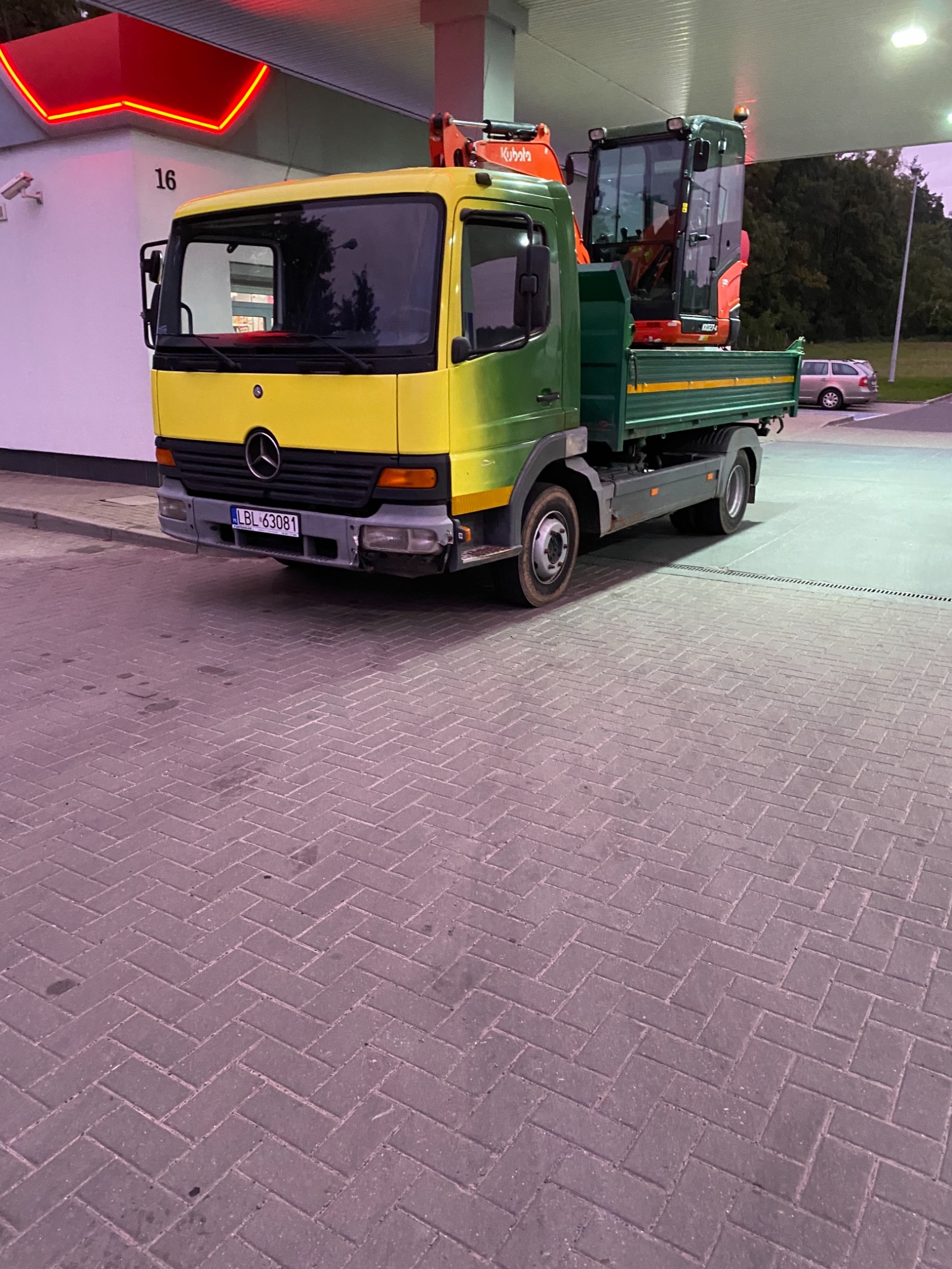 Żółto-zielona ciężarówka Mercedes z pomarańczową mini-koparką Kubota na pace, zaparkowana przy budynku z neonowym numerem 16.
