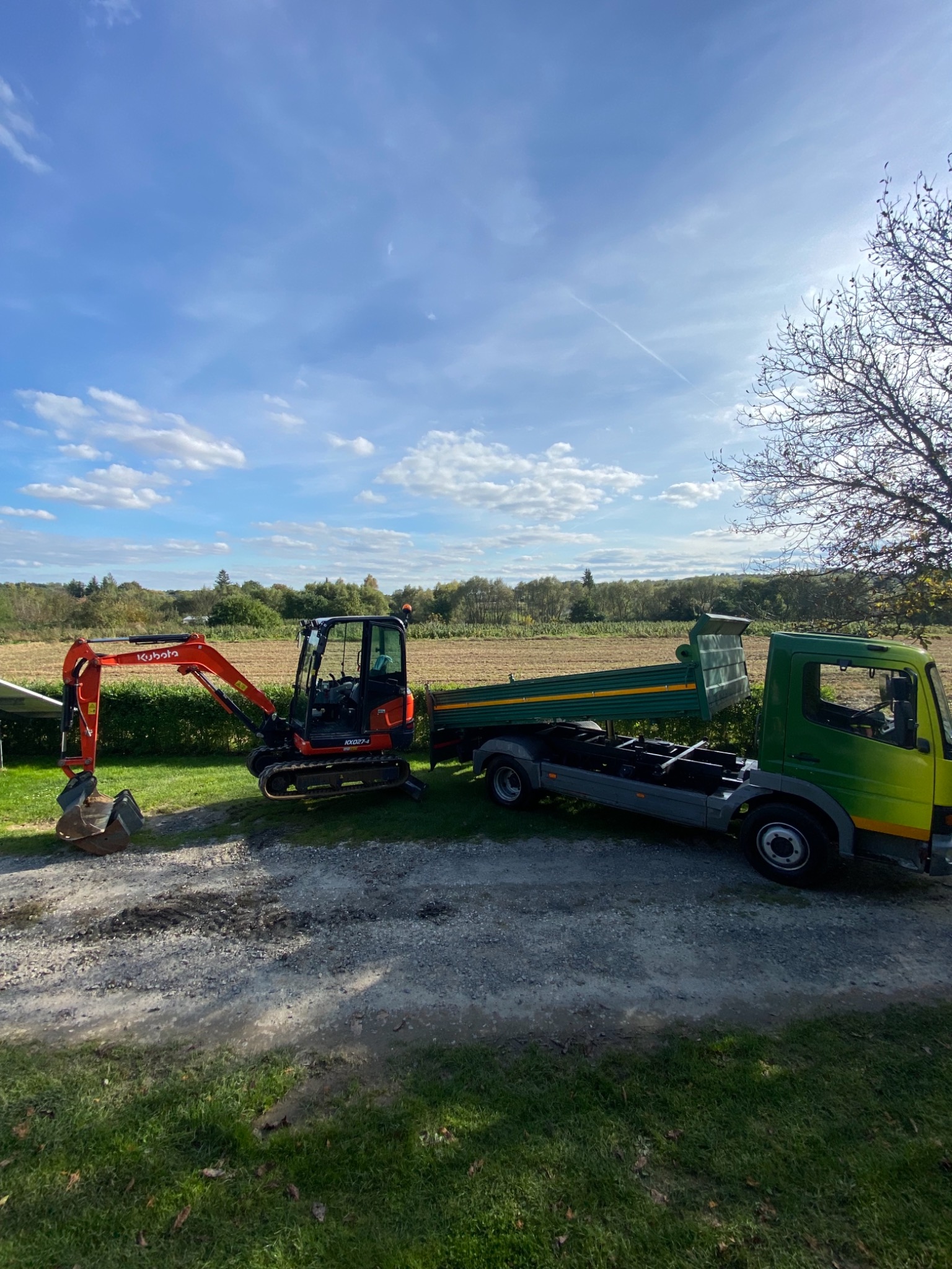 Pomarańczowa minikoparka Kubota obok zielono-żółtej wywrotki na żwirowej drodze, w tle pole i błękitne niebo z chmurami.