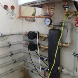 Fragment kotłowni z widocznymi miedzianymi i plastikowymi rurami, manometrem, dwoma pompami obiegowymi i przewodami elektrycznymi wystającymi ze ściany.