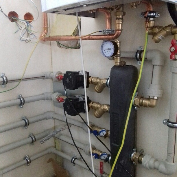 Fragment kotłowni z widocznymi miedzianymi i plastikowymi rurami, manometrem, dwoma pompami obiegowymi i przewodami elektrycznymi wystającymi ze ściany.