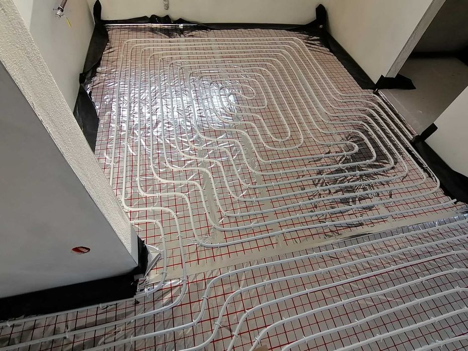 Białe rurki ogrzewania podłogowego ułożone na folii z czerwoną siatką, widoczne fragmenty ścian w surowym stanie.