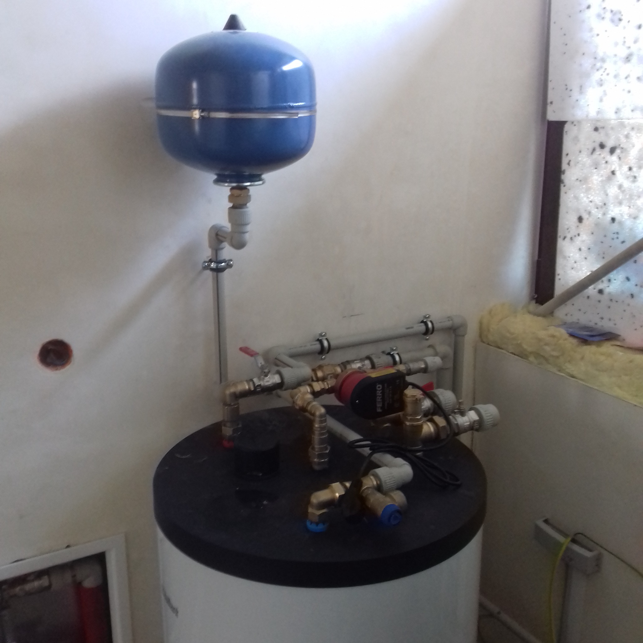 Niebieski zasobnik ciśnieniowy zamontowany nad białym zbiornikiem z podłączeniami hydraulicznymi i pompą obiegową Ferroli, widoczne rury i armatura, ściana z niedoskonałościami i śladami po...