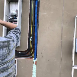 Instalacja elektryczna w ścianie zewnętrznej budynku, widoczne kable w peszlach, licznik elektryczny w skrzynce oraz pianka montażowa uszczelniająca przejścia przez mur.