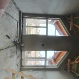 Wnętrze poddasza z dwoma oknami dachowymi po nałożeniu gładzi, widoczne drewniane elementy konstrukcji dachu oraz wiadro i narzędzia na podłodze.