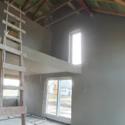 Wnętrze domu w trakcie prac wykończeniowych: ściany pokryte świeżym tynkiem, widoczna drewniana drabina, okna z widokiem na sąsiedni budynek, instalacje elektryczne wystające ze ścian.