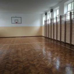 Sala gimnastyczna z parkietem w jodełkę, drabinki gimnastyczne przy ścianie i kosz do koszykówki, po odświeżeniu koloru ścian.