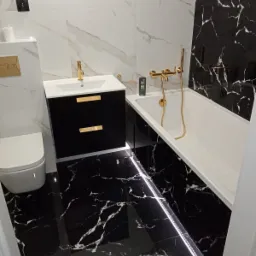 Elegancka łazienka z czarno-białymi marmurkowymi płytkami, złotą armaturą, podświetleniem LED pod wanną i czarną szafką umywalkową z złotymi uchwytami.