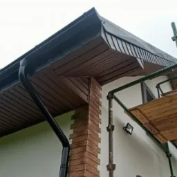 Fragment elewacji domu z drewnianą podbitką, czarną rynną i rusztowaniem w tle, widok z dołu.