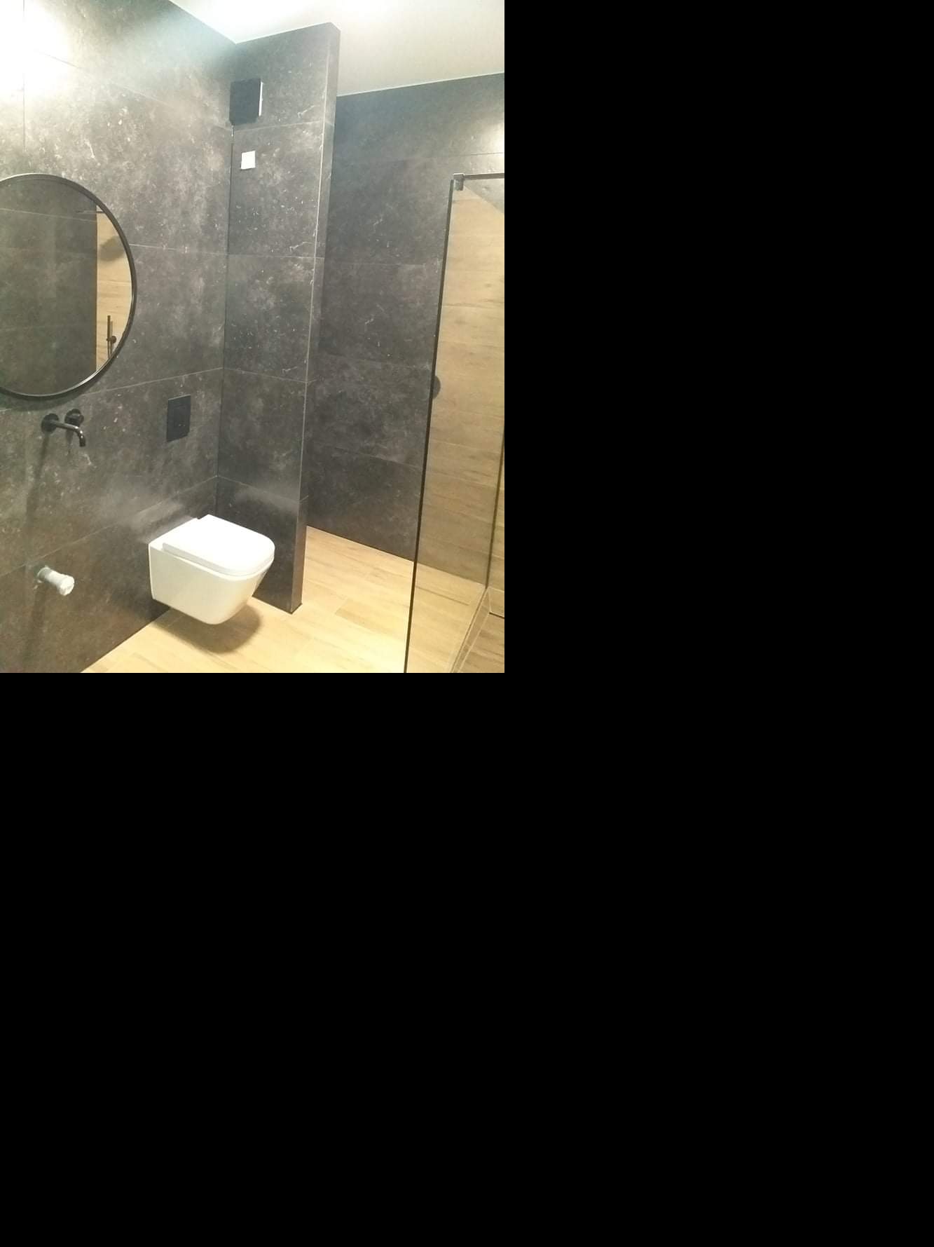 Nowoczesna łazienka z szarymi płytkami imitującymi kamień, okrągłe lustro, podwieszana toaleta i kabina prysznicowa z drewnopodobną podłogą.