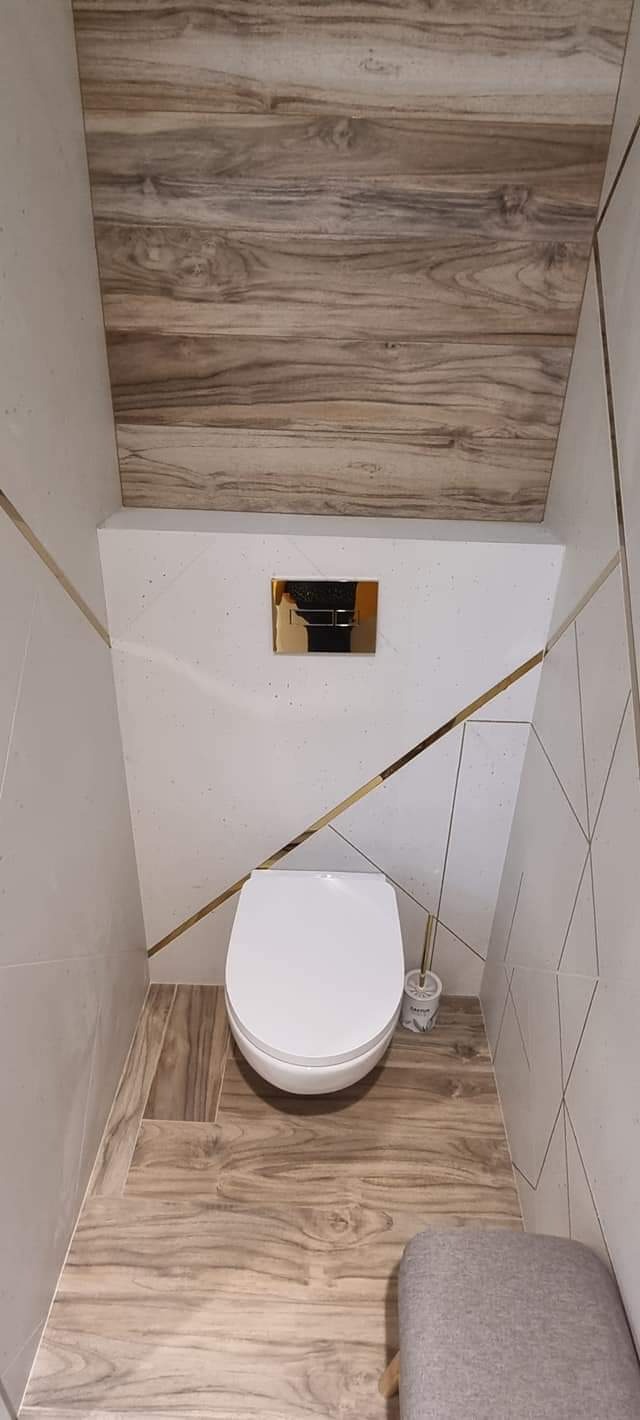 Nowoczesna toaleta w łazience z drewnopodobnymi płytkami na podłodze i suficie, białe ściany z geometrycznym wzorem podkreślonym złotymi listwami, wisząca miska WC i szczotka do WC.