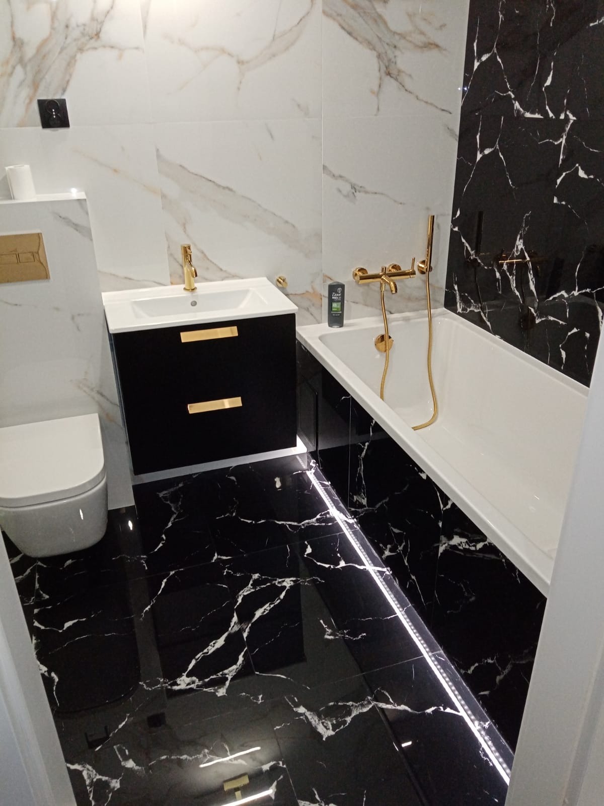 Elegancka łazienka z czarno-białymi marmurkowymi płytkami, złotą armaturą, podświetleniem LED pod wanną i czarną szafką umywalkową z złotymi uchwytami.