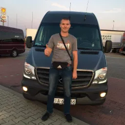 Mężczyzna z uniesionym kciukiem stoi przed ciemnym busem marki Mercedes-Benz Sprinter na tle stacji benzynowej Shell i restauracji Burger King.