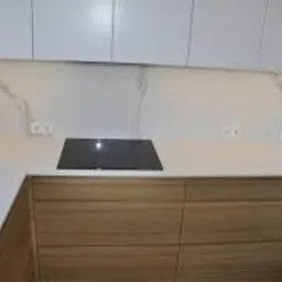 Blat kuchenny z jasnego konglomeratu z wbudowaną czarną płytą indukcyjną, podświetlony punktowo, z widocznymi gniazdkami elektrycznymi na ścianie.