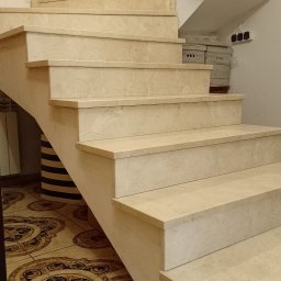 Usługi Kamieniarskie Marcin Mrozek - Kremowe, spiralne schody z kamienia z widocznym wzorem na stopniach i posadzce, z grzejnikiem i niezamaskowanymi gniazdkami elektrycznymi w tle.