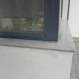 Usługi Kamieniarskie Marcin Mrozek - Szary parapet zewnętrzny z konglomeratu pod oknem z moskitierą, widoczna kostka brukowa na zewnątrz.