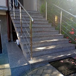 Usługi Kamieniarskie Marcin Mrozek - Zewnętrzne schody z szarego kamienia z metalową balustradą, prowadzące do wejścia domu, z widoczną wycieraczką i kwiatami w tle.