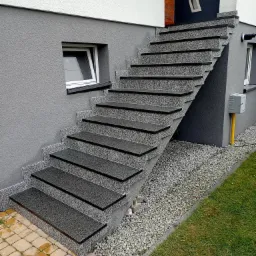 Zewnętrzne schody wejściowe z szarego kamienia z czarnymi krawędziami, prowadzące do ciemnych drzwi wejściowych w nowoczesnym domu z szarą elewacją i białym pasem na g&oacute;rze, otoczone jasnym żwirem...