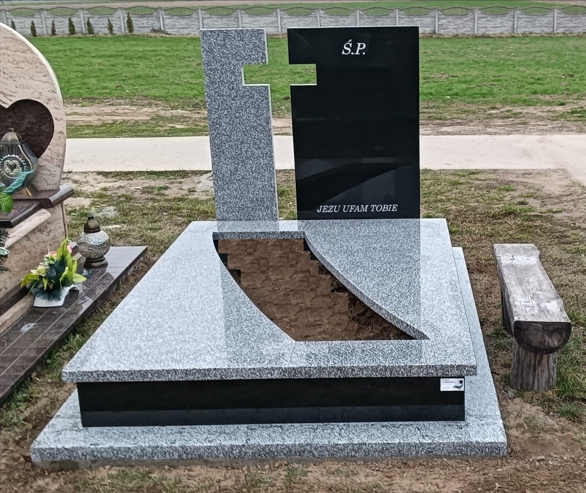 Nowoczesny nagrobek z jasnego i czarnego kamienia, z wyciętym fragmentem płyty i napisem 'Jezu ufam Tobie', na tle zielonego trawnika.