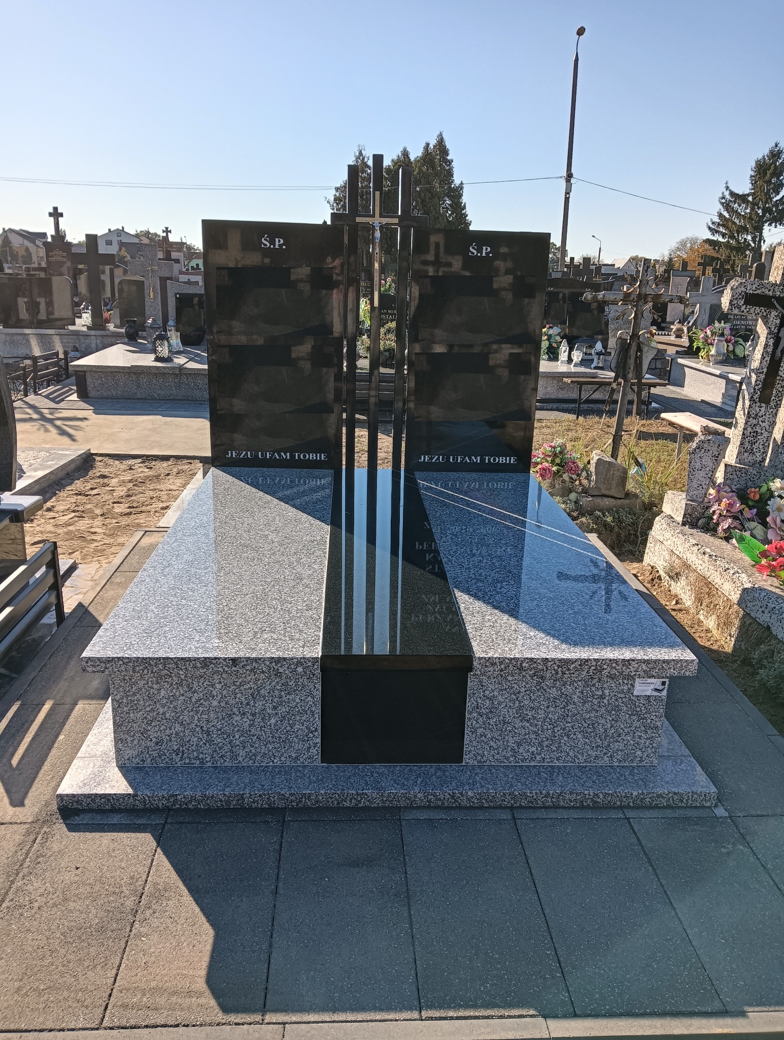 Nowoczesny nagrobek z ciemnego granitu i jasnoszarego kamienia, z metalowym krzyżem, na tle cmentarza w słoneczny dzień. Napis: 'Jezu ufam Tobie'.