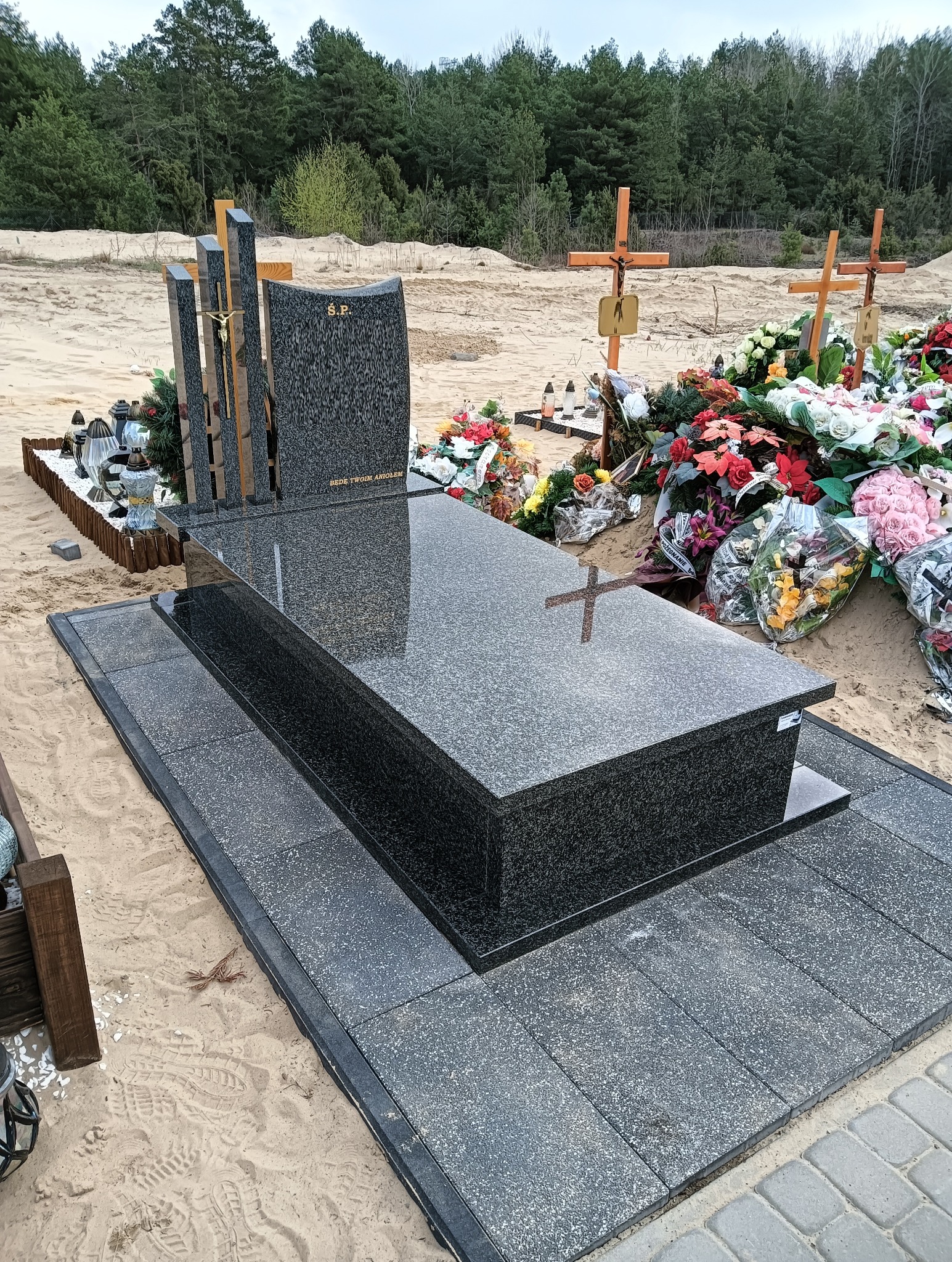 Granitowy nagrobek z krzyżem, otoczony kwiatami i zniczami na piaszczystym terenie cmentarza, w tle drzewa. Widoczne detale wykończenia i wysoka jakość kamienia.