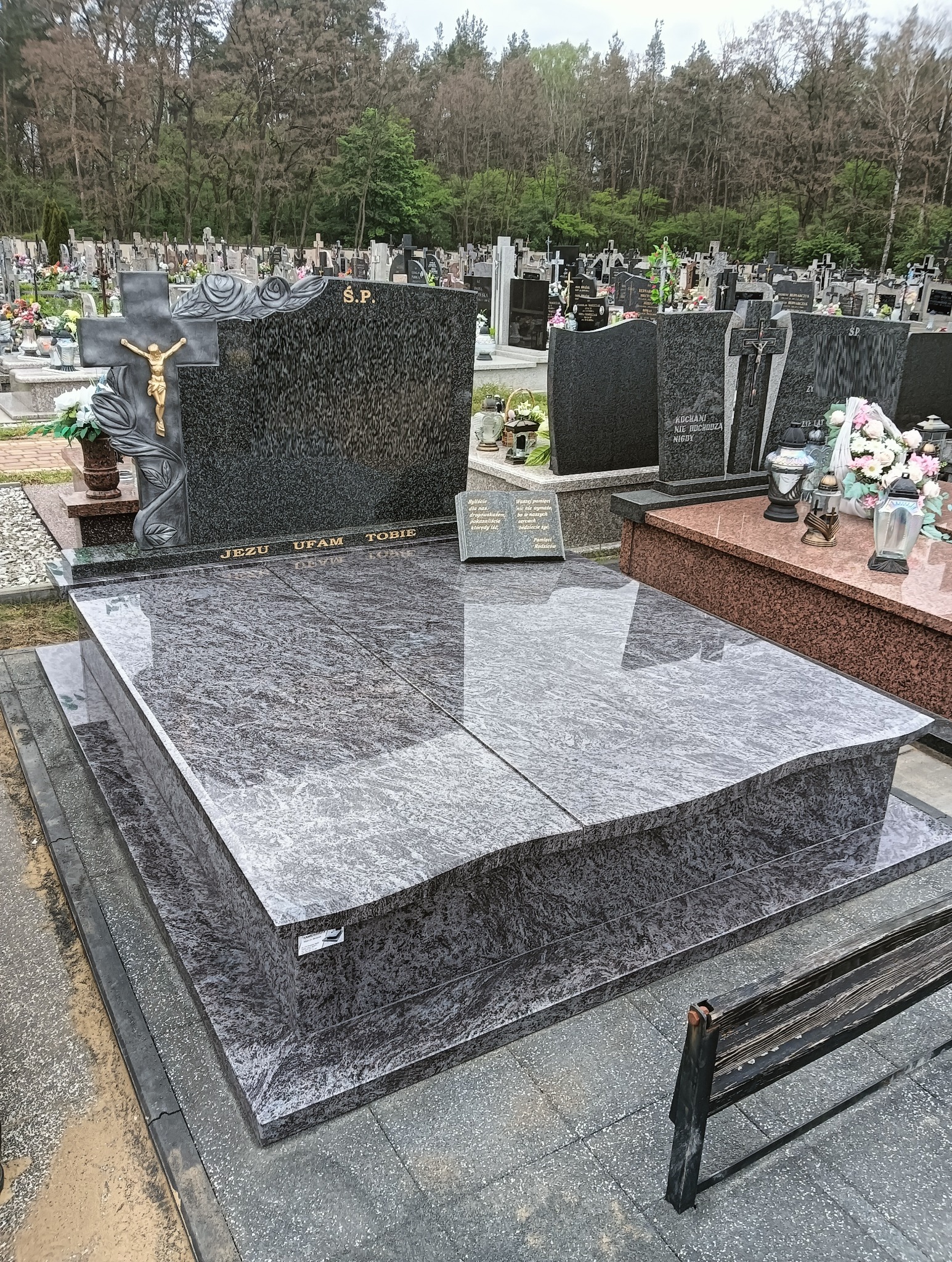 Granitowy nagrobek z krzyżem i inskrypcją 'Jezu Ufam Tobie' na cmentarzu, z widocznymi nagrobkami w tle i fragmentem ławki. Kamień polerowany.